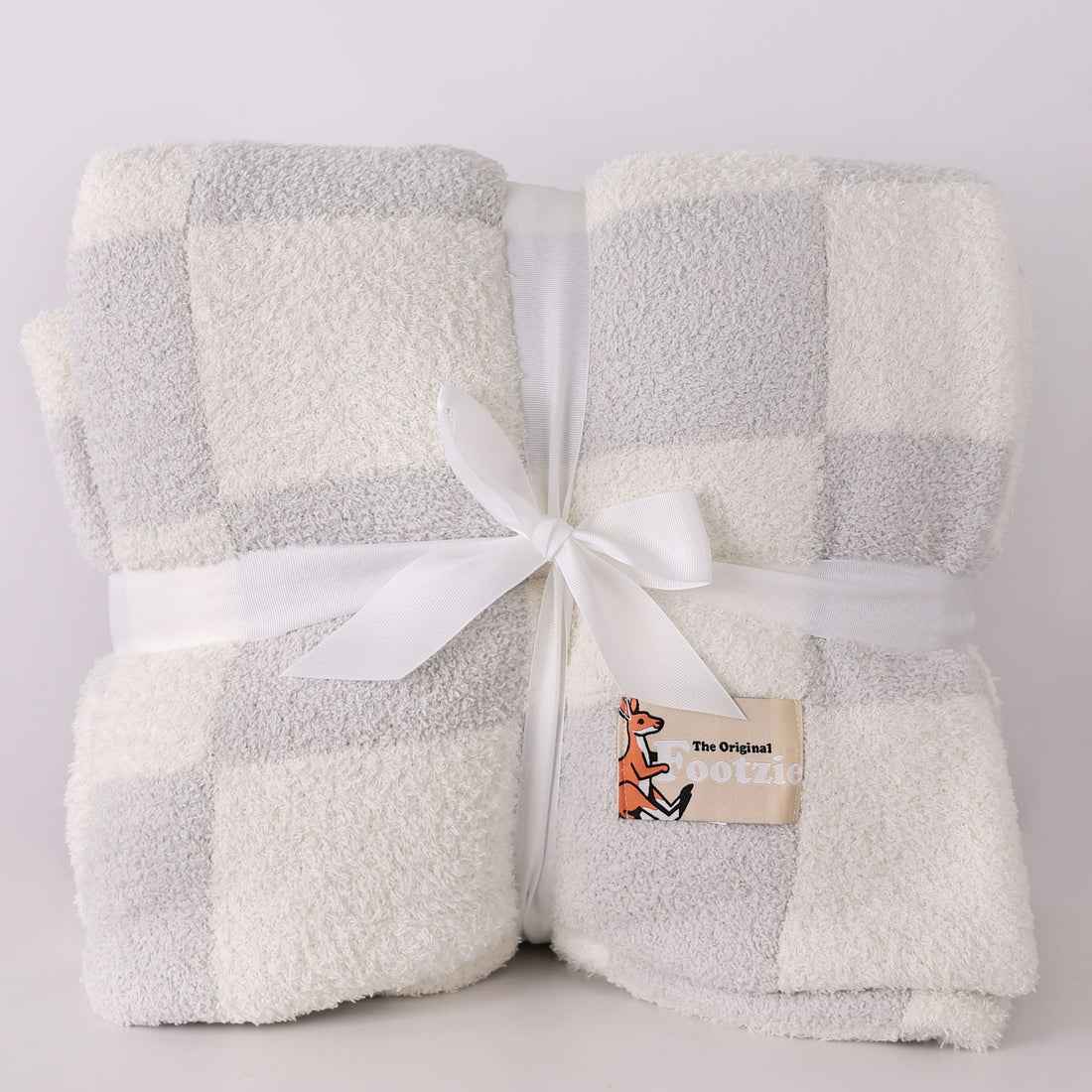 Folded checkered blanket with a white ribbon and a brand label on a light gray background, blanket has a pouch for your feet