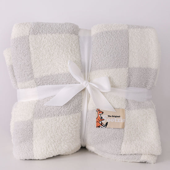 Folded checkered blanket with a white ribbon and a brand label on a light gray background, blanket has a pouch for your feet